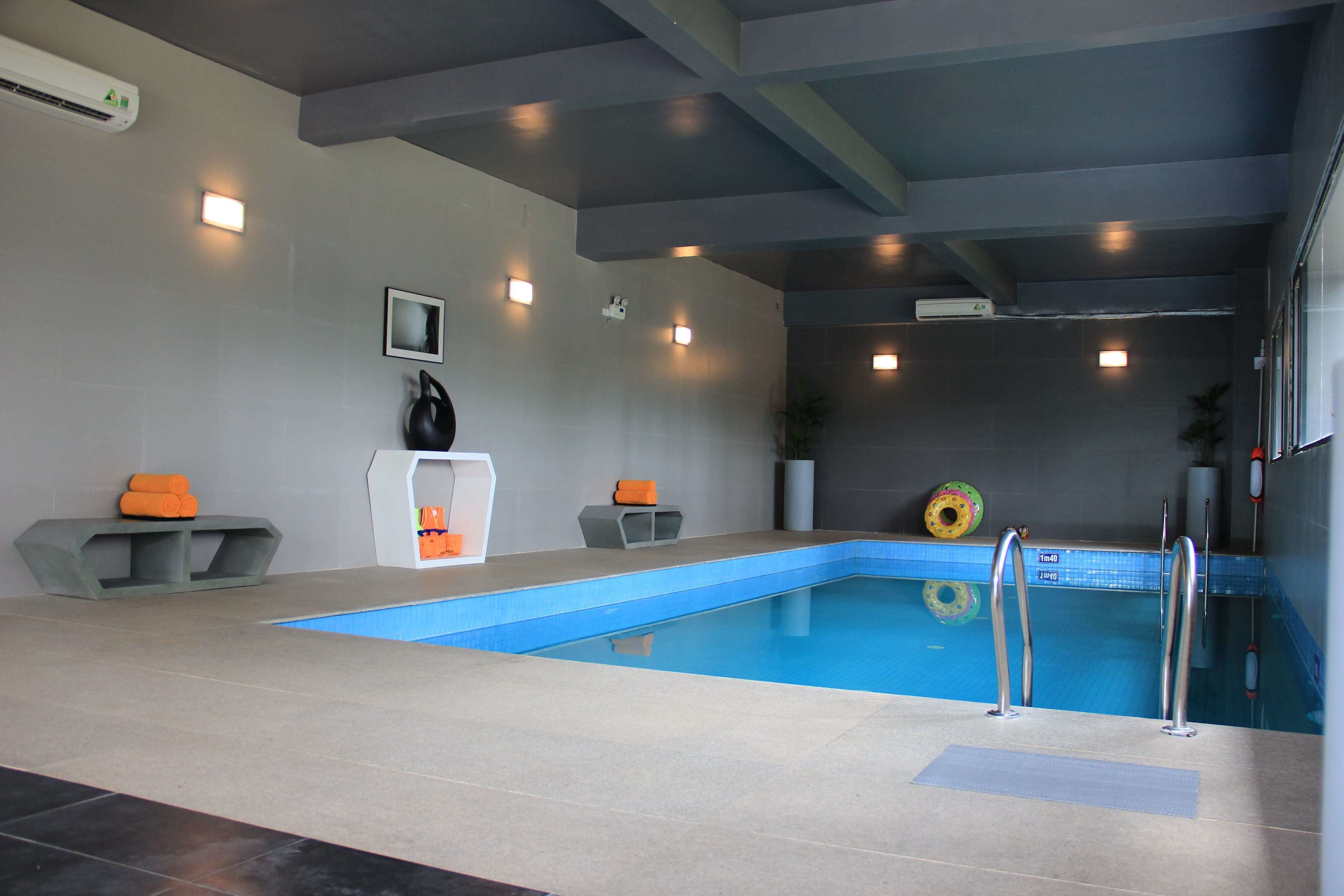A swimming pool with a blue interior, surrounded by grey walls and a ceiling with exposed beams. There are two small seating areas with orange cushions, a white pedestal, and a framed picture on the wall.