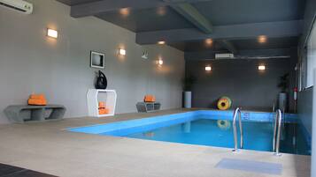 A swimming pool with a blue interior, surrounded by grey walls and a ceiling with exposed beams. There are two small seating areas with orange cushions, a white pedestal, and a framed picture on the wall.