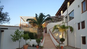 A stone-paved courtyard with a staircase leading to a wooden deck, surrounded by potted plants and a white building with multiple windows. - B&B Mediterraneo Mare e Sole (Palma di Montechiaro)