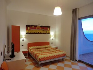 A hotel room with a bed, a desk, a chair, a TV, a painting on the wall, and a window with curtains. - B&B Mediterraneo Mare e Sole (Palma di Montechiaro)