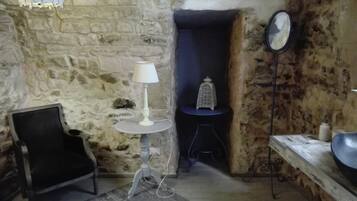A rustic room with a stone wall, a white lamp on a small table, a black chair, a small stool, a lantern, and a round mirror on a stand.