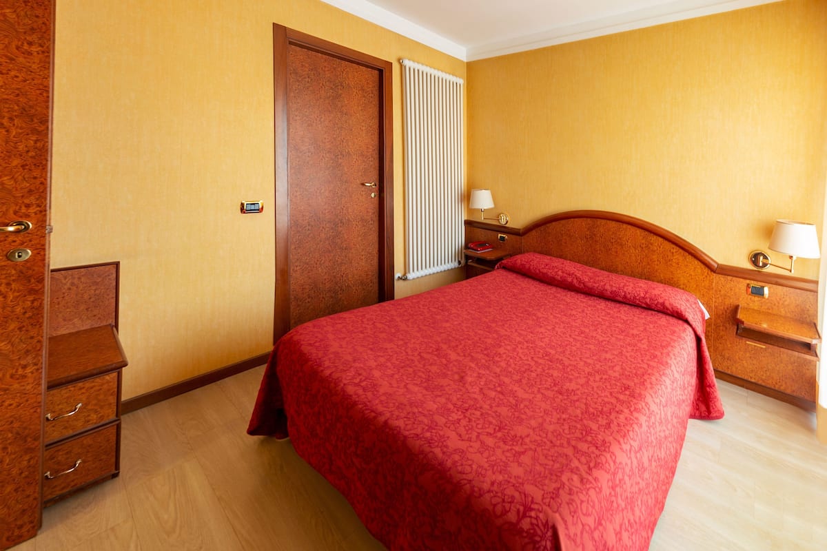 Una habitación de hotel con una cama, un armario de madera, una mesita de noche, una lámpara y una ventana con persianas.