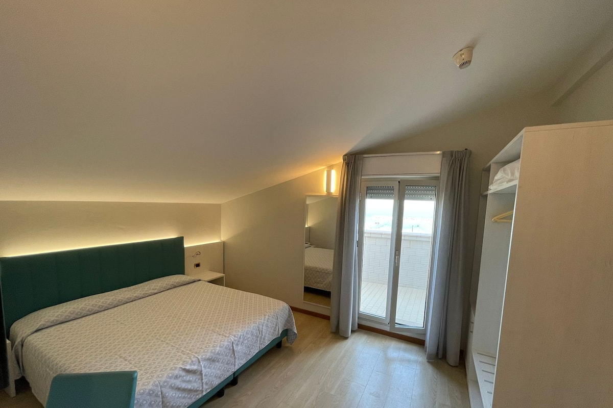 Habitación de hotel con cama, escritorio, silla, armario y puerta corrediza de vidrio al balcón.