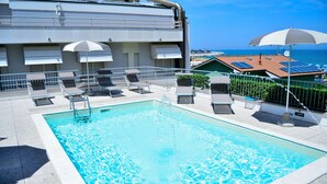 Une piscine sur le toit, dotée de chaises longues et de parasols, offrant une vue sur une zone côtière avec des bâtiments et la mer.