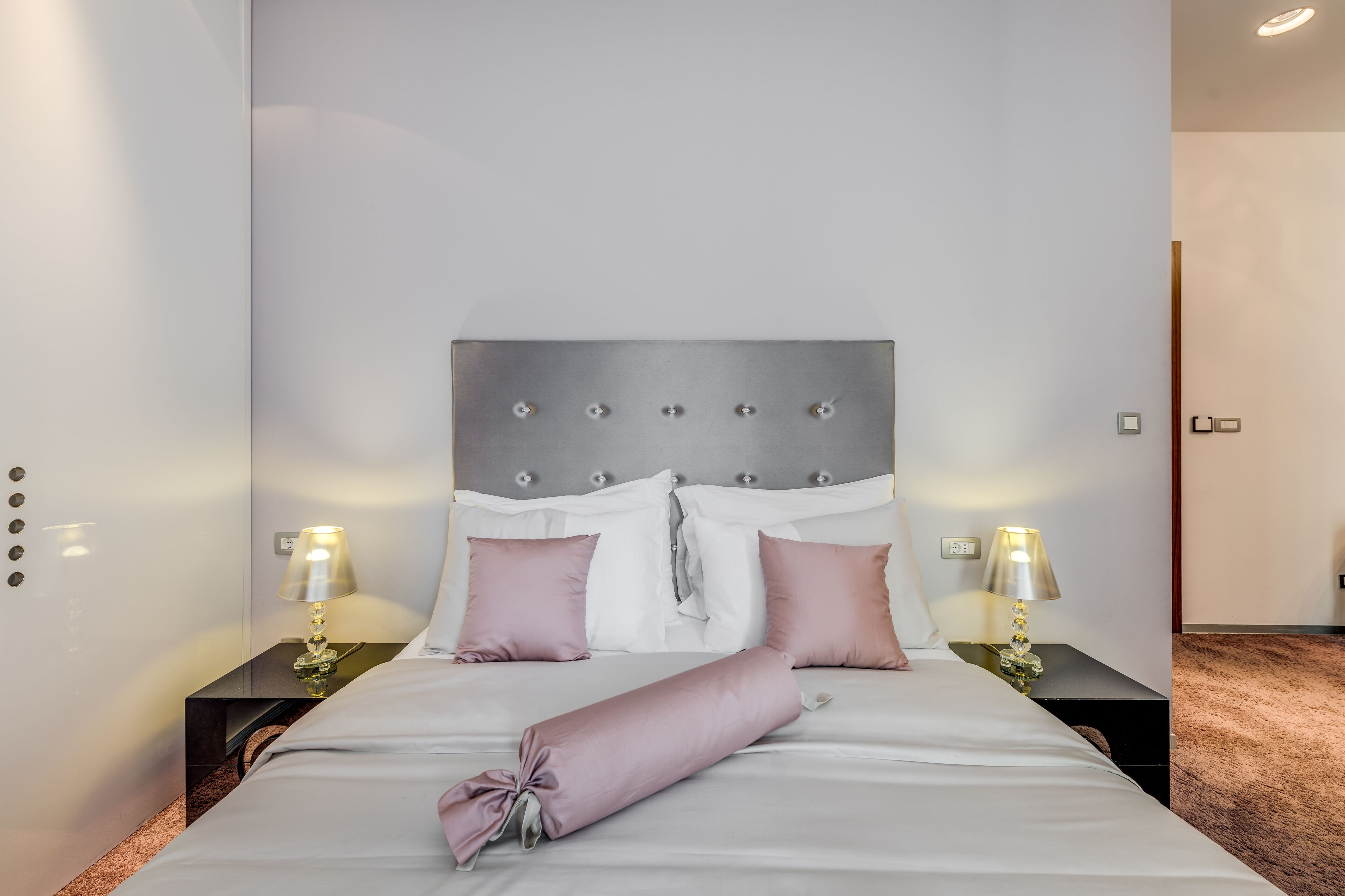 A neatly made bed with a grey headboard, two bedside tables with lamps, and a carpeted floor.