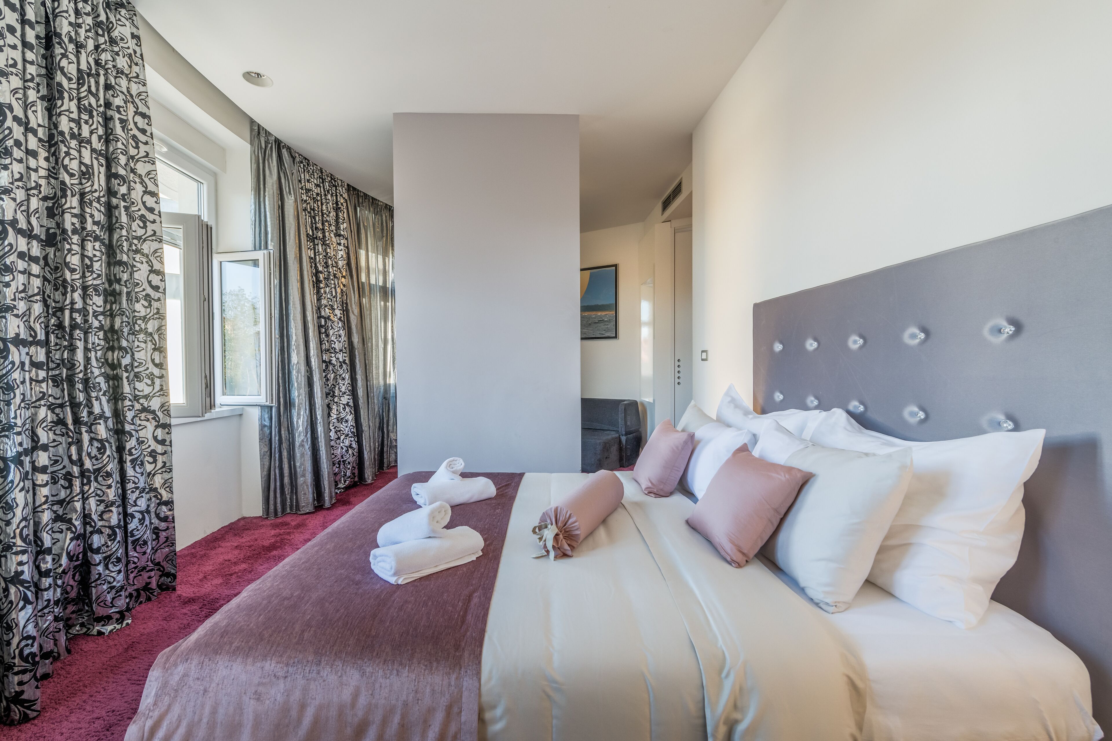 A hotel room with a large bed, a red carpet, and a view of the outdoors through the window.