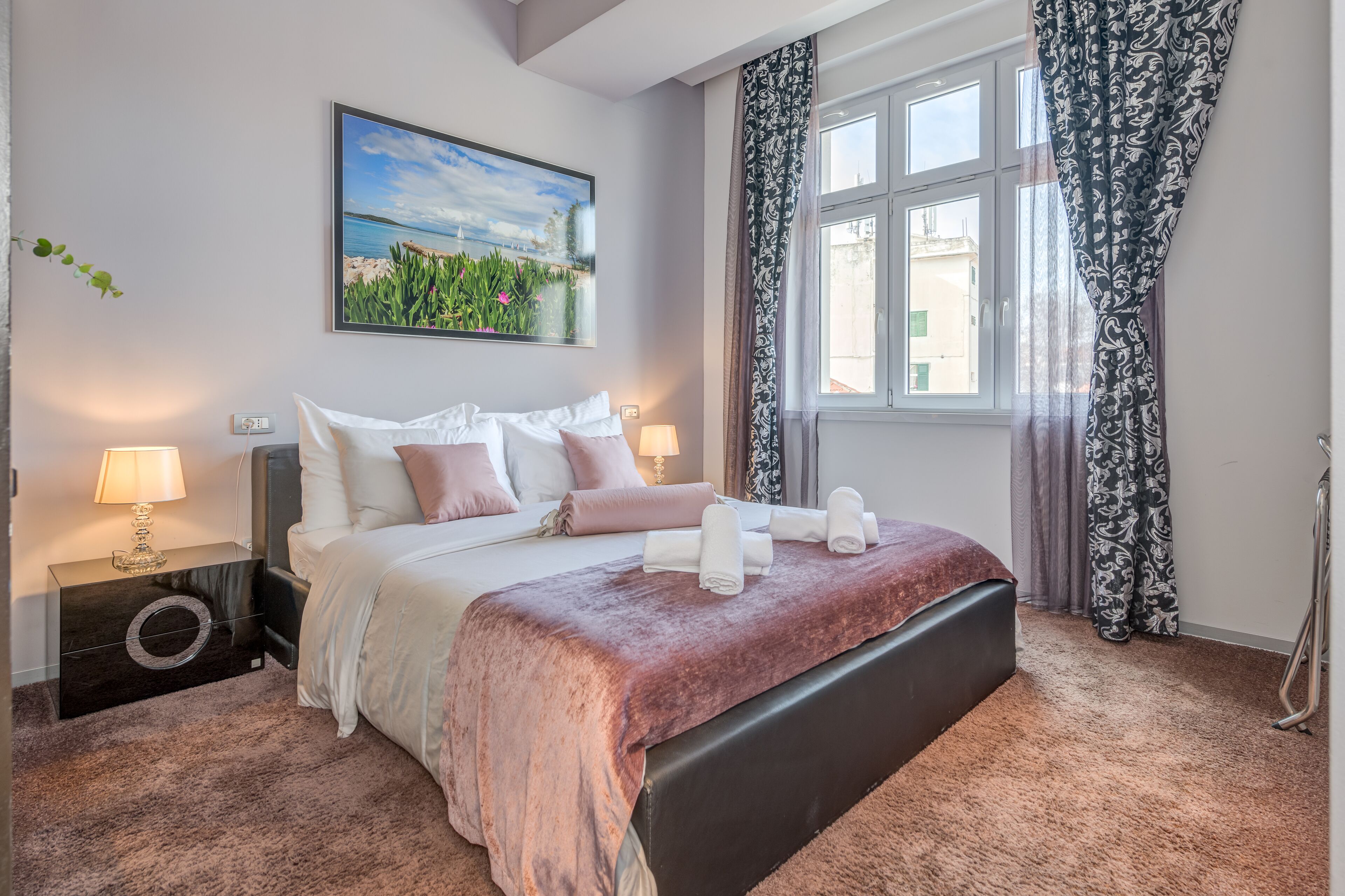 Een slaapkamer met een bed, nachtlampen, een nachtkastje, een raam met gordijnen en een schilderij aan de muur.
