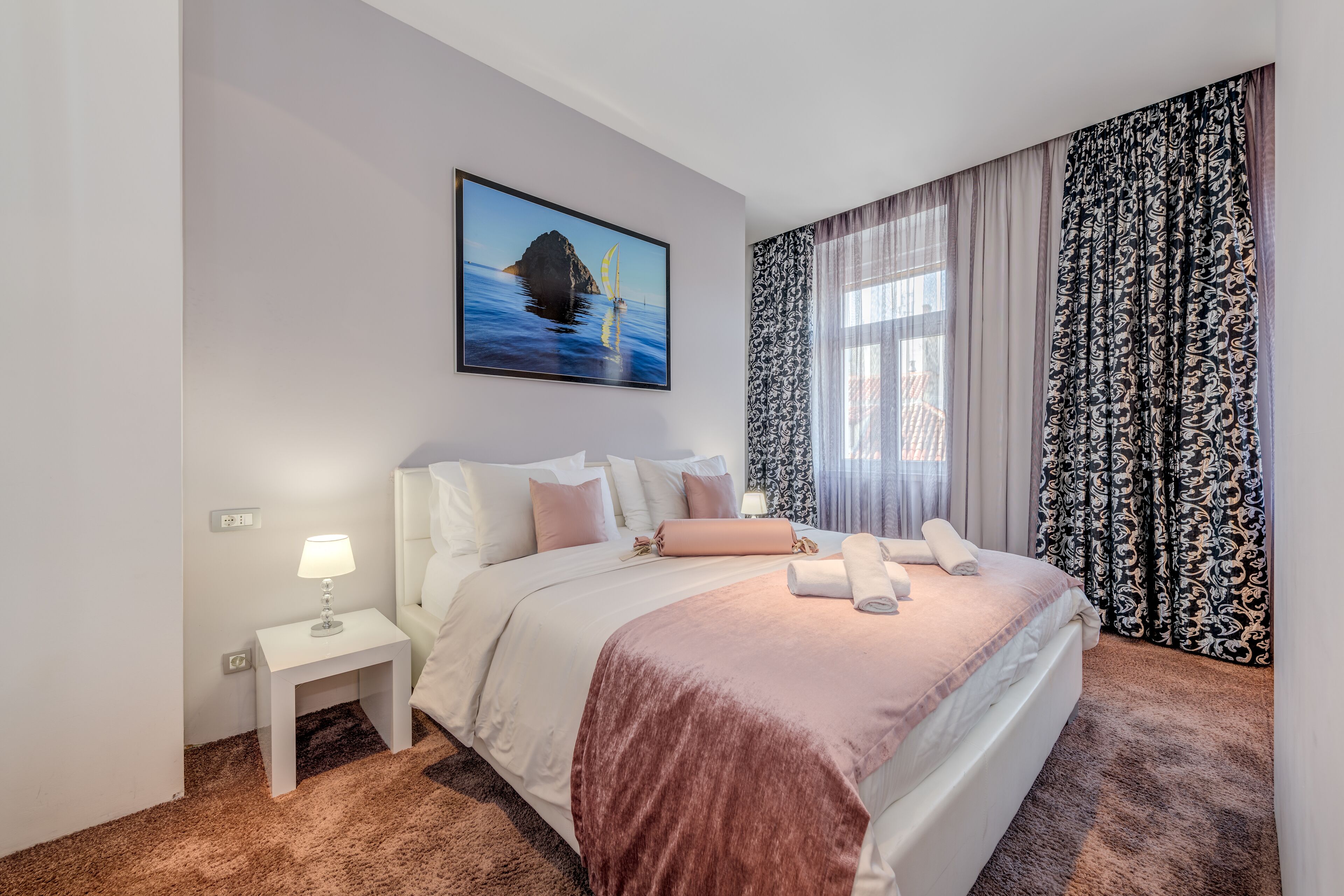A hotel room with a large bed, a nightstand with a lamp, a painting of a sailboat, and patterned curtains.