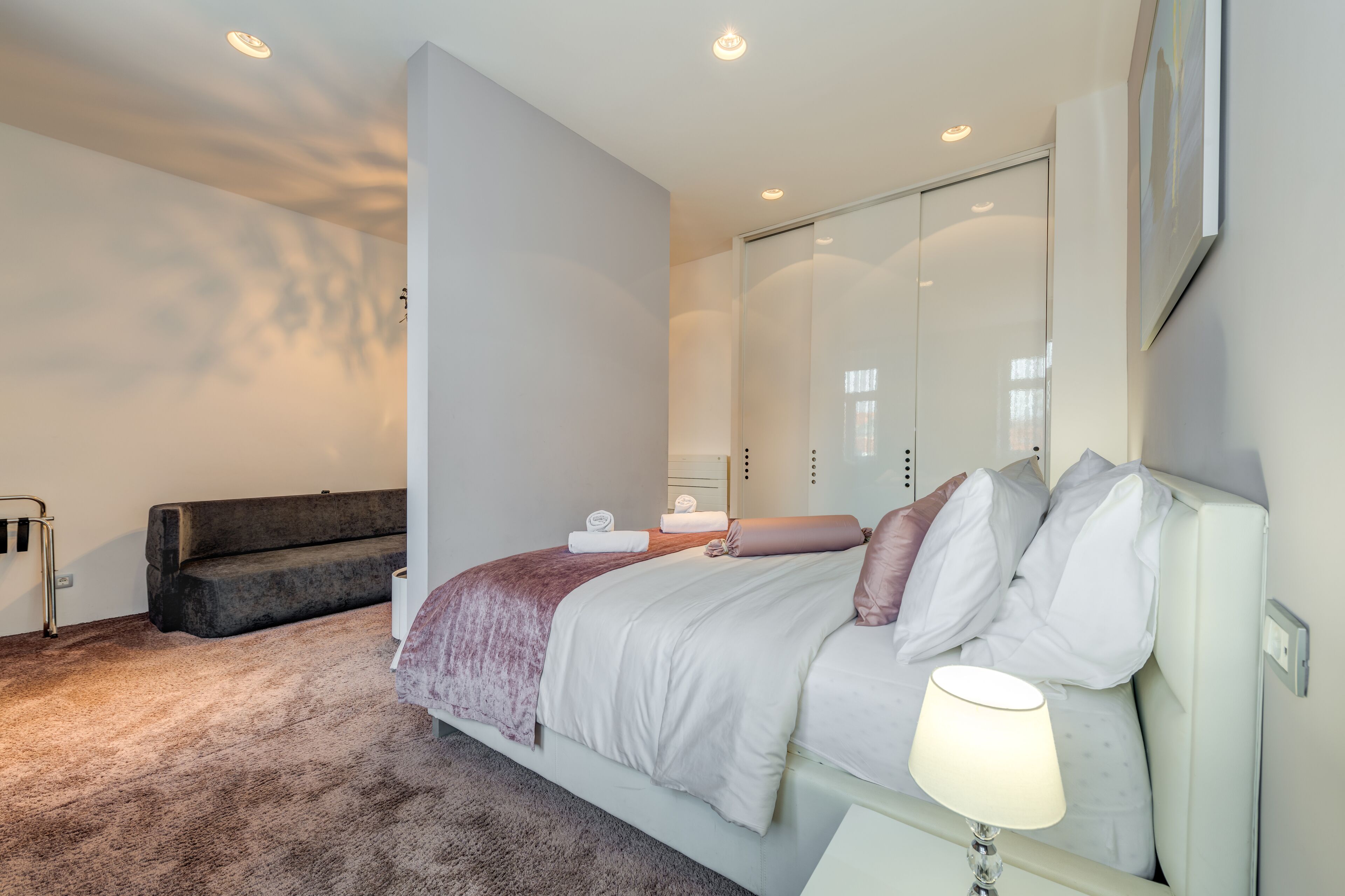 A modern bedroom with a large bed, a bedside table with a lamp, a dark sofa, and a walk-in closet.