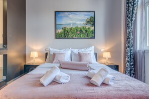 Ett hotellrum med en stor säng, två sänglampor, en tavla som föreställer en strandscen och utsikt över havet genom fönstret.