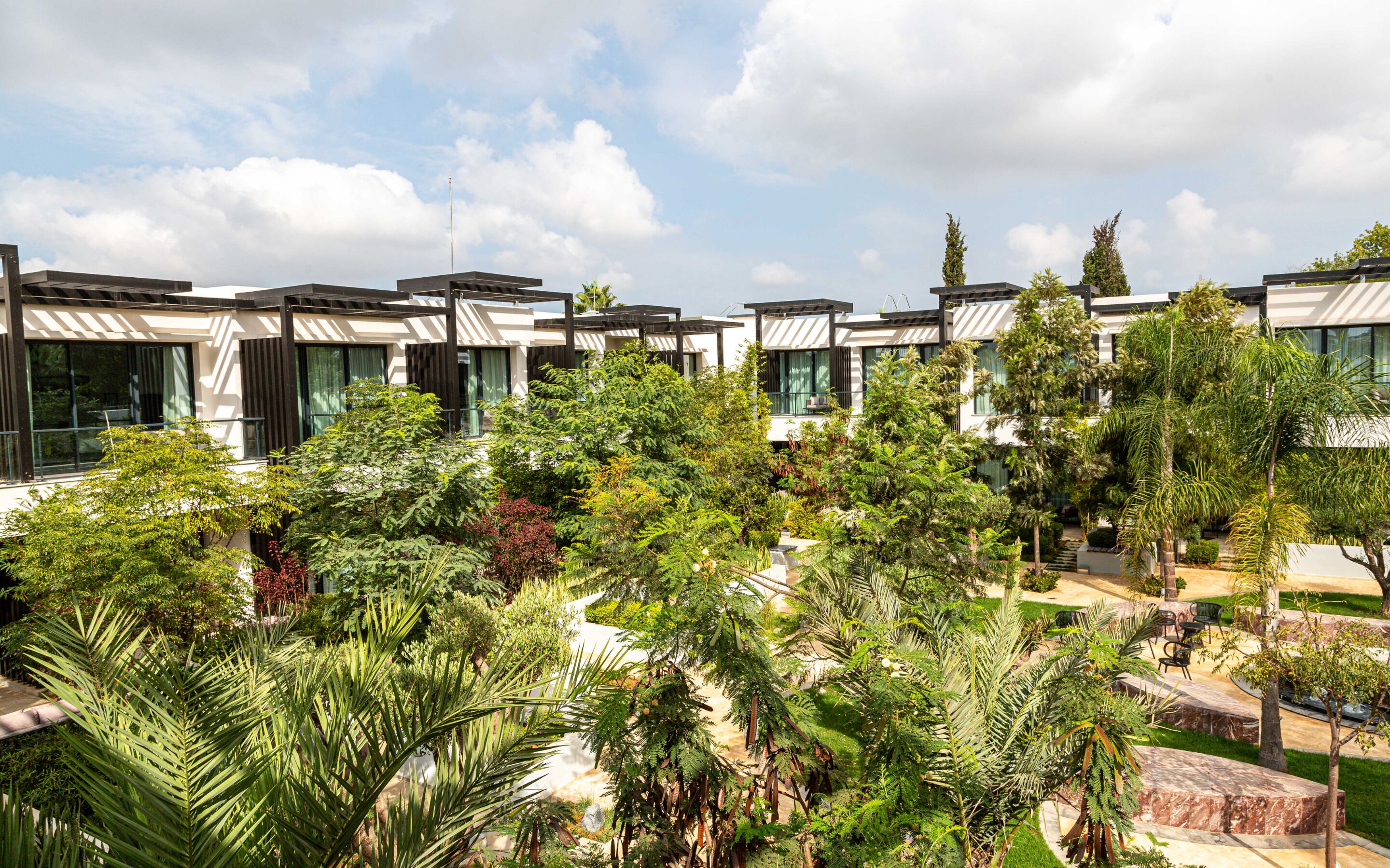 A modern hotel with multiple-story buildings, lush greenery, and a clear sky.