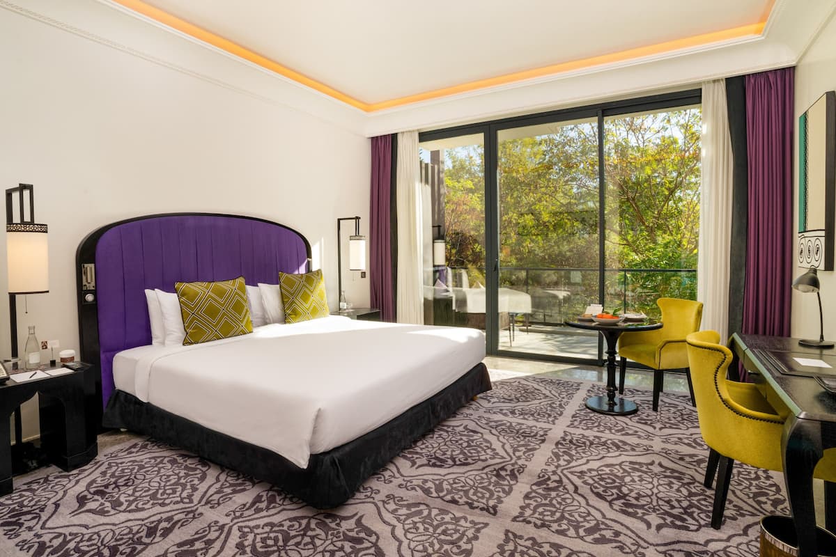A hotel room with a large bed, a desk, a chair, a balcony with a table, and a view of trees.