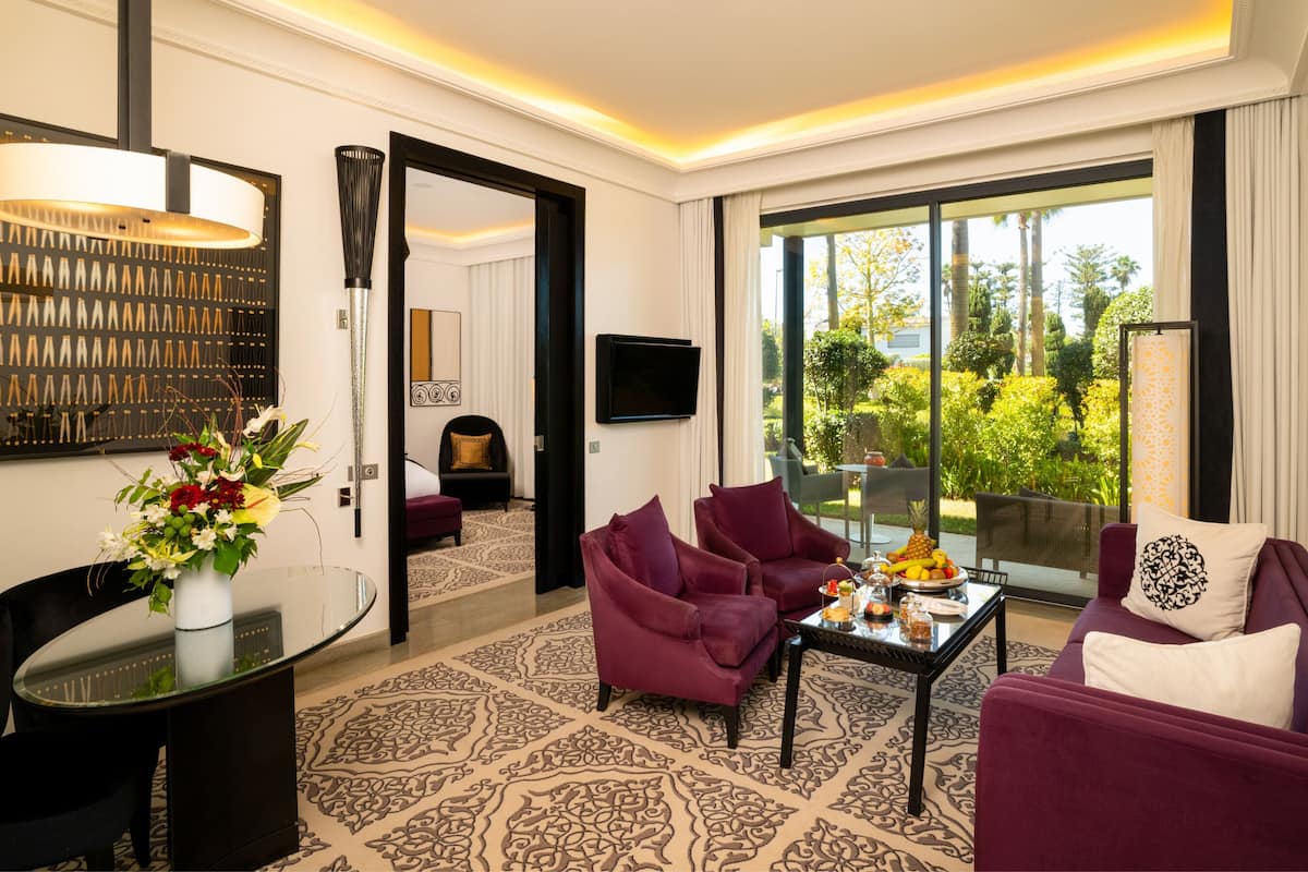 A modern hotel room with a large window, a flat-screen TV, a dining table, and two purple armchairs.