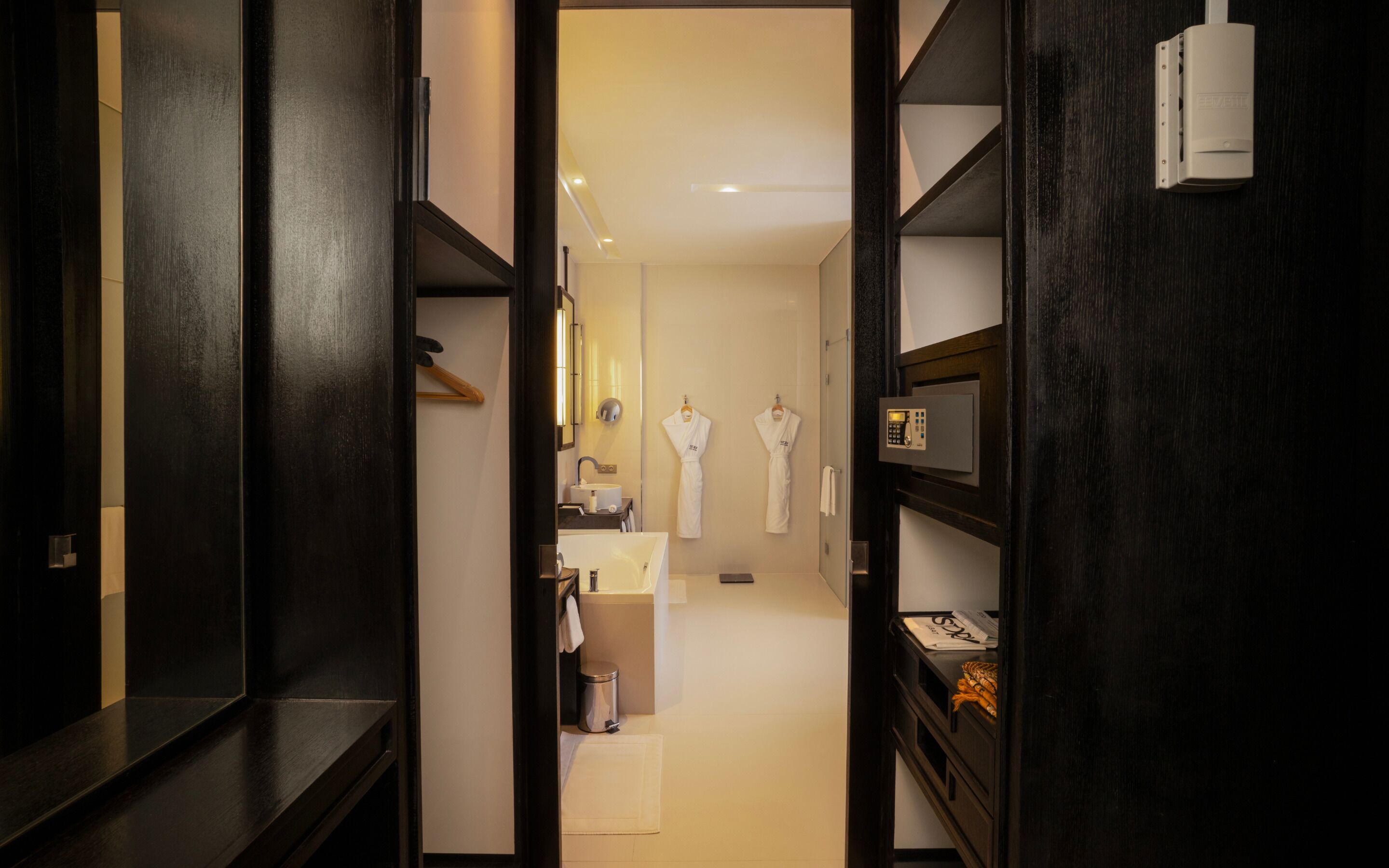 Un couloir d’hôtel avec un miroir, un lavabo et deux peignoirs blancs accrochés au mur.