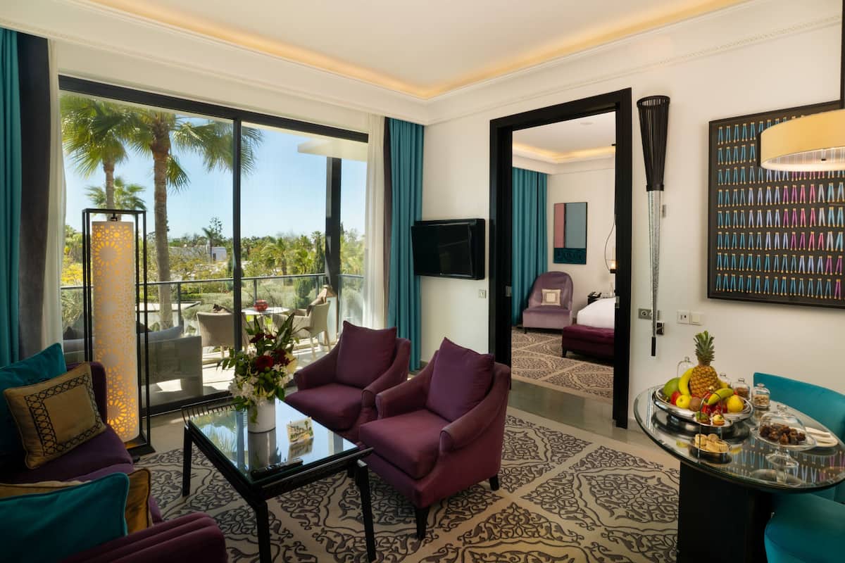 A hotel room with a balcony, a TV, a dining area with a fruit bowl, and a patterned carpet.