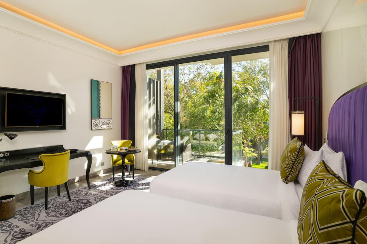 A hotel room with a bed, a desk, a chair, a TV, and a balcony with a view of trees.