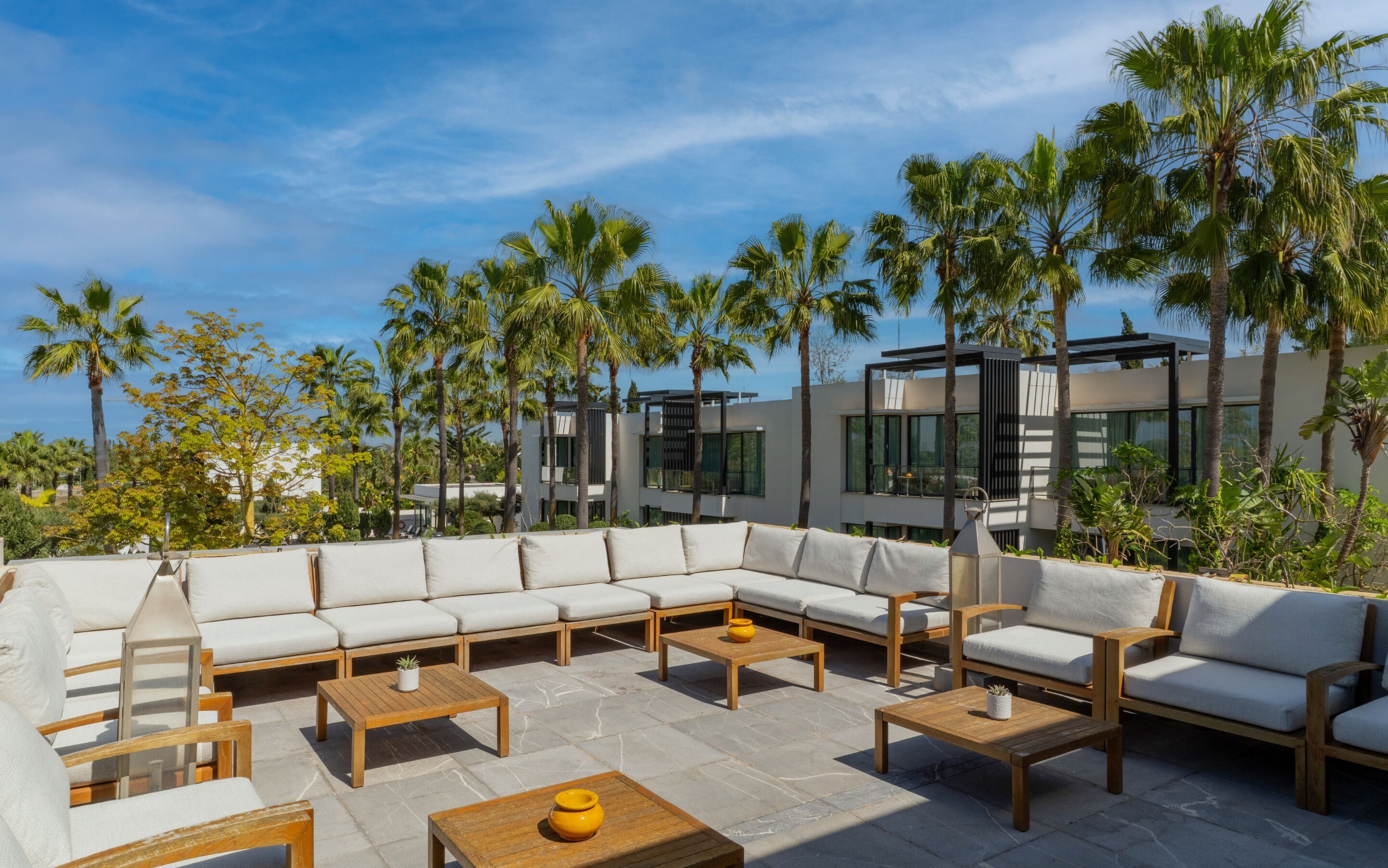 A modern outdoor seating area with a sectional sofa, wooden tables, and a view of a contemporary building with large windows.