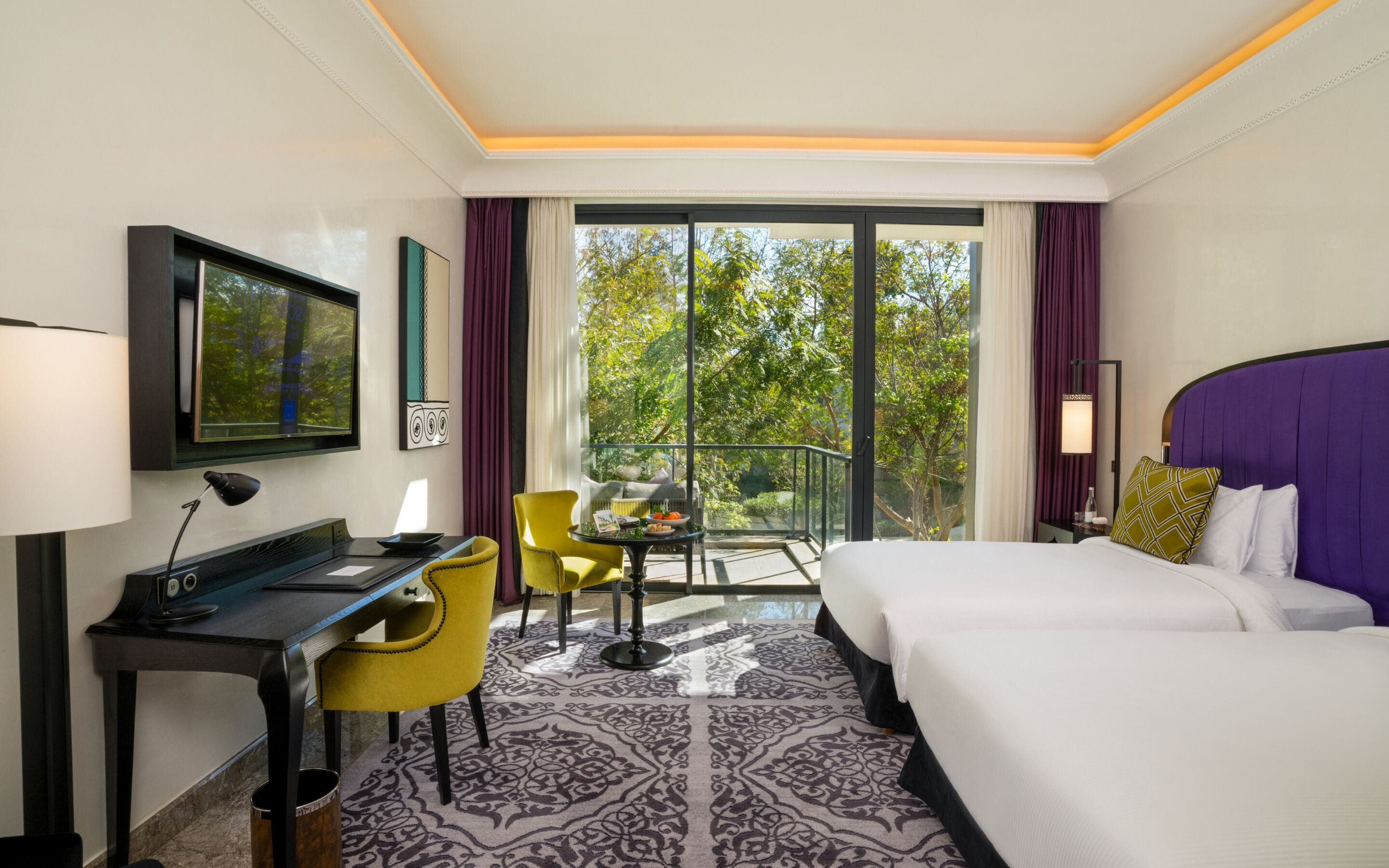 A hotel room with a bed, desk, chair, and a balcony with a view of trees.