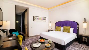 Una habitación de hotel con una cama, un escritorio con una silla, una mesita con un frutero y una mesita de noche con una lámpara.