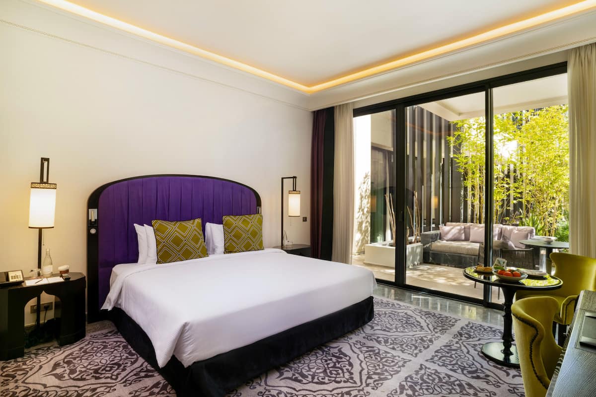 A hotel room with a large bed, a desk with a lamp, a chair, and a view of greenery outside.