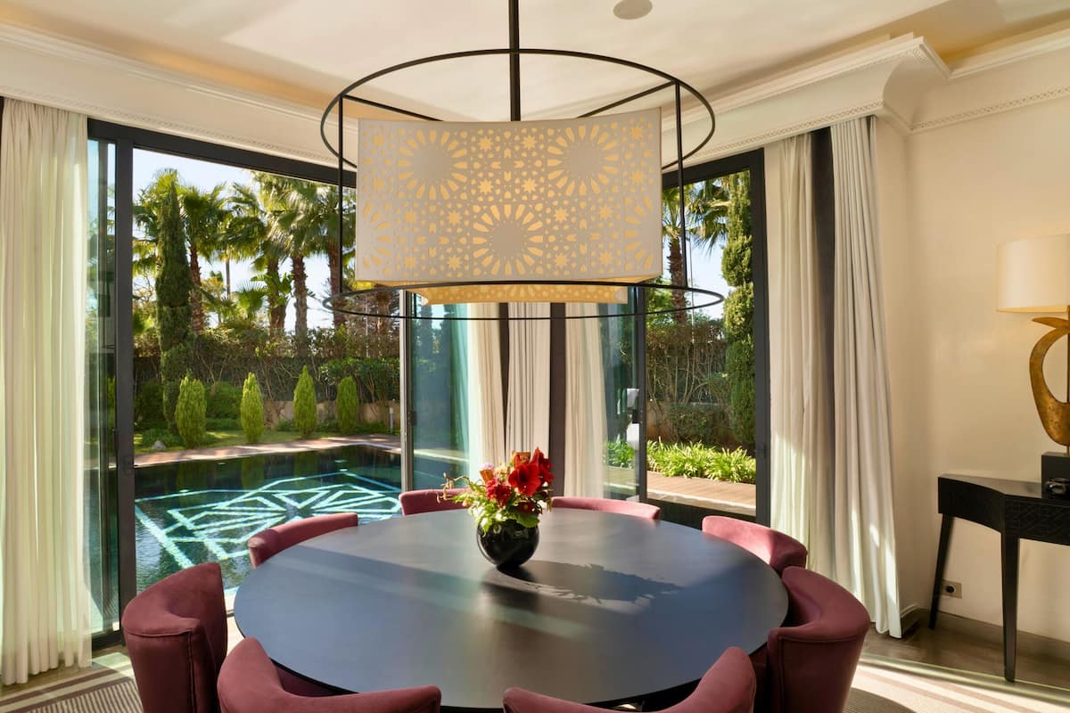 A modern room with a round table, red chairs, a decorative lamp, and a view of a pool and greenery.