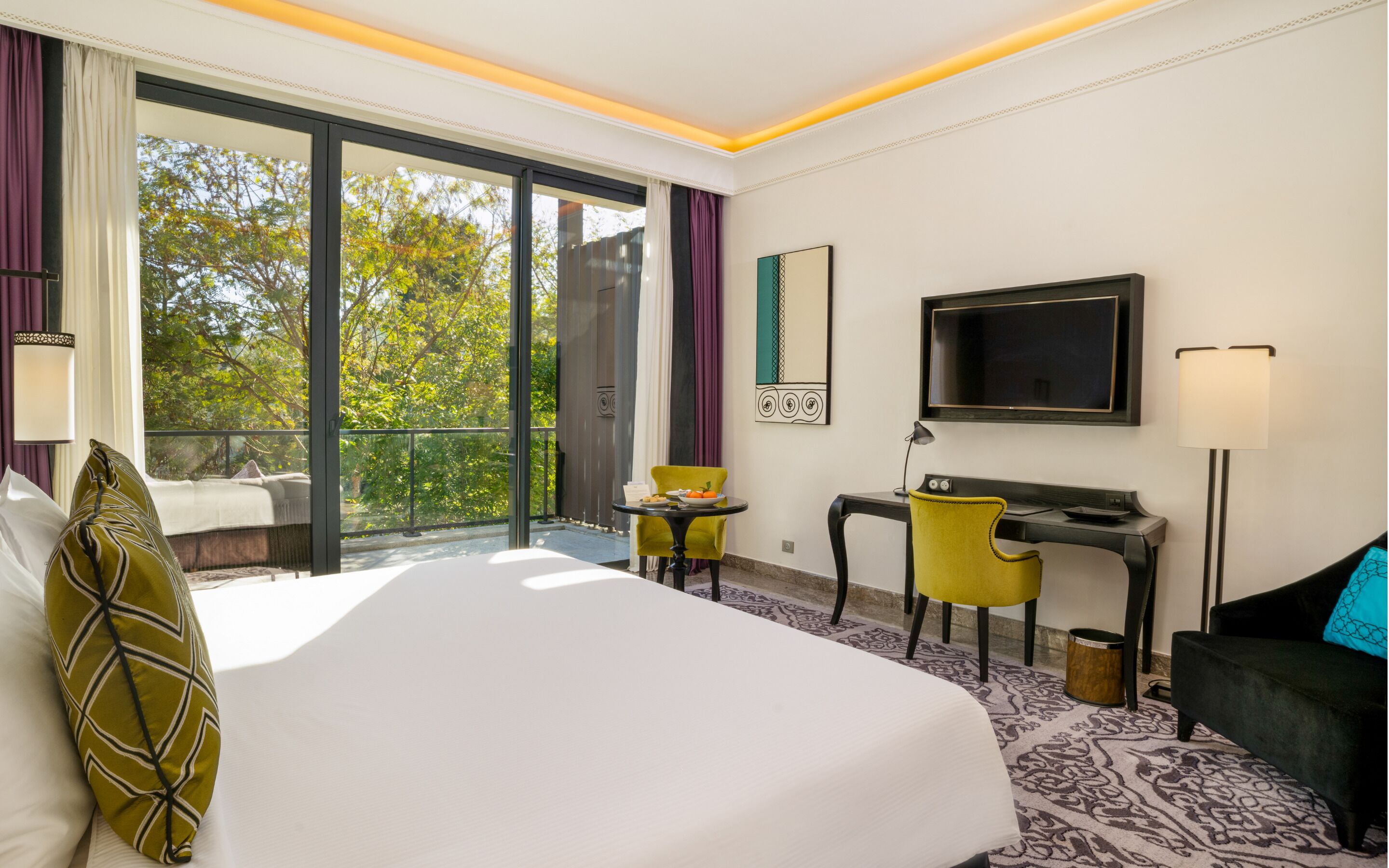 A hotel room with a bed, desk, chair, TV, and a balcony with a view of trees.
