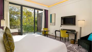 Quarto de hotel com cama, escrivaninha, cadeira, televisão e varanda com vista para árvores.