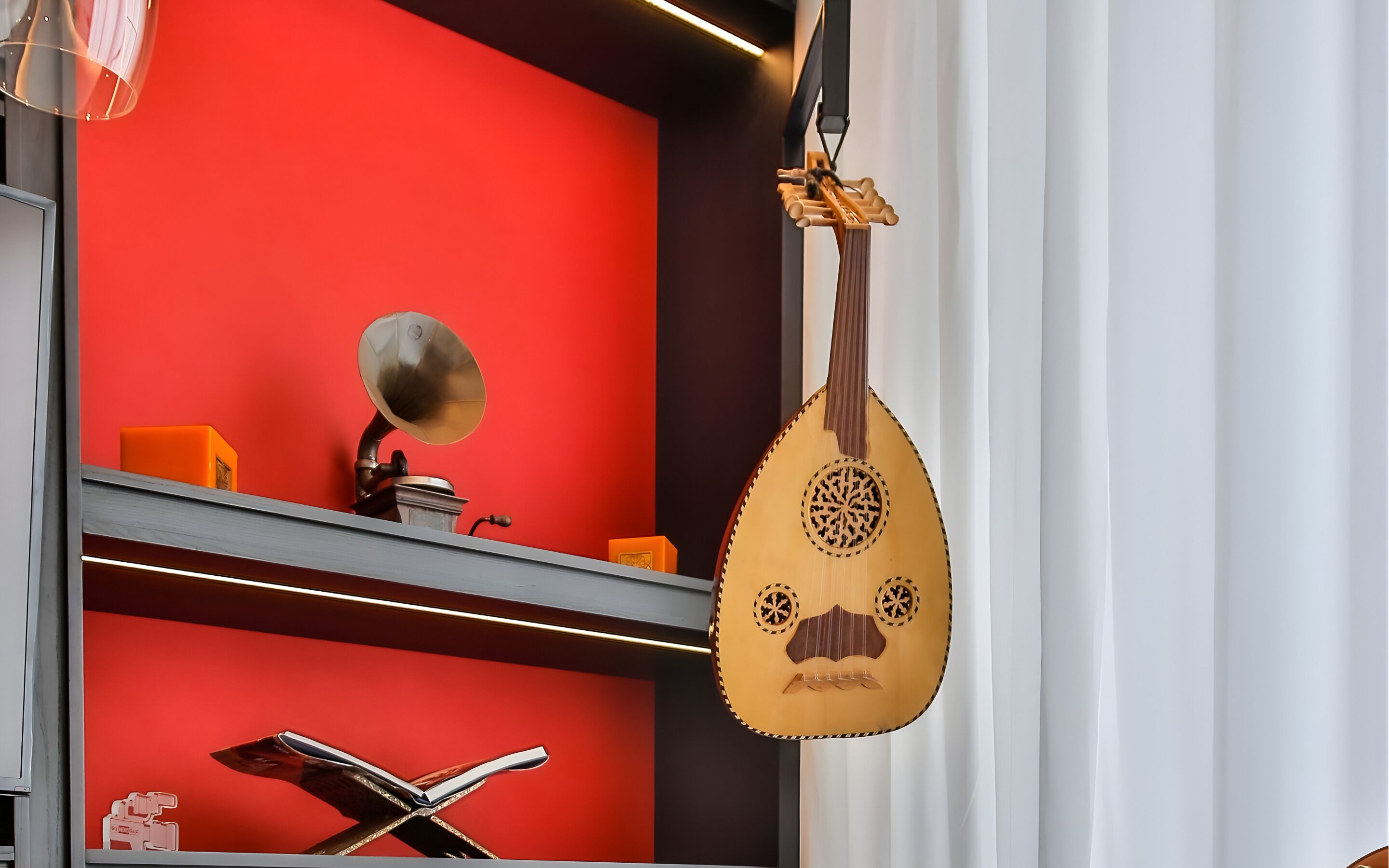 Ein Zimmer mit roter Wand, einem Grammophon, einem hängenden Saiteninstrument und einem Regal mit Büchern und einer kleinen orangefarbenen Schachtel.