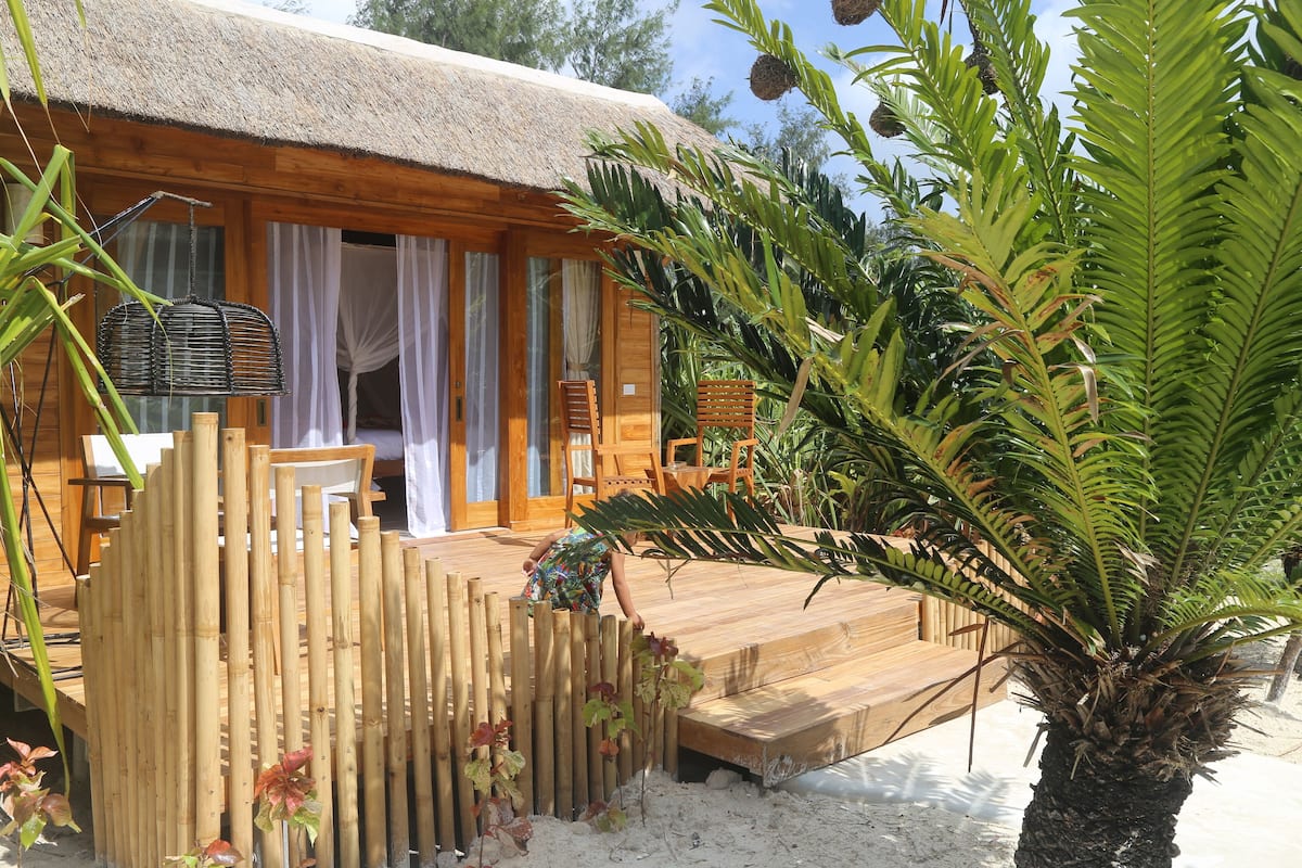 A wooden cabin with a thatched roof, a wooden deck, and a palm tree.