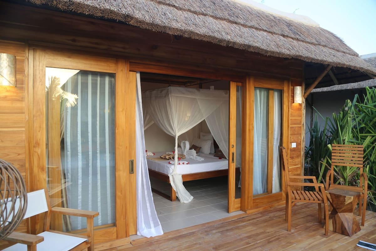 A wooden room with a bed, curtains, and a thatched roof.
