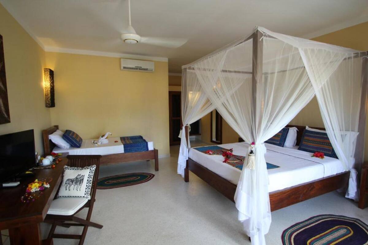 A bedroom with a four-poster bed, a desk with a chair, a television, and a bedside table with a lamp.