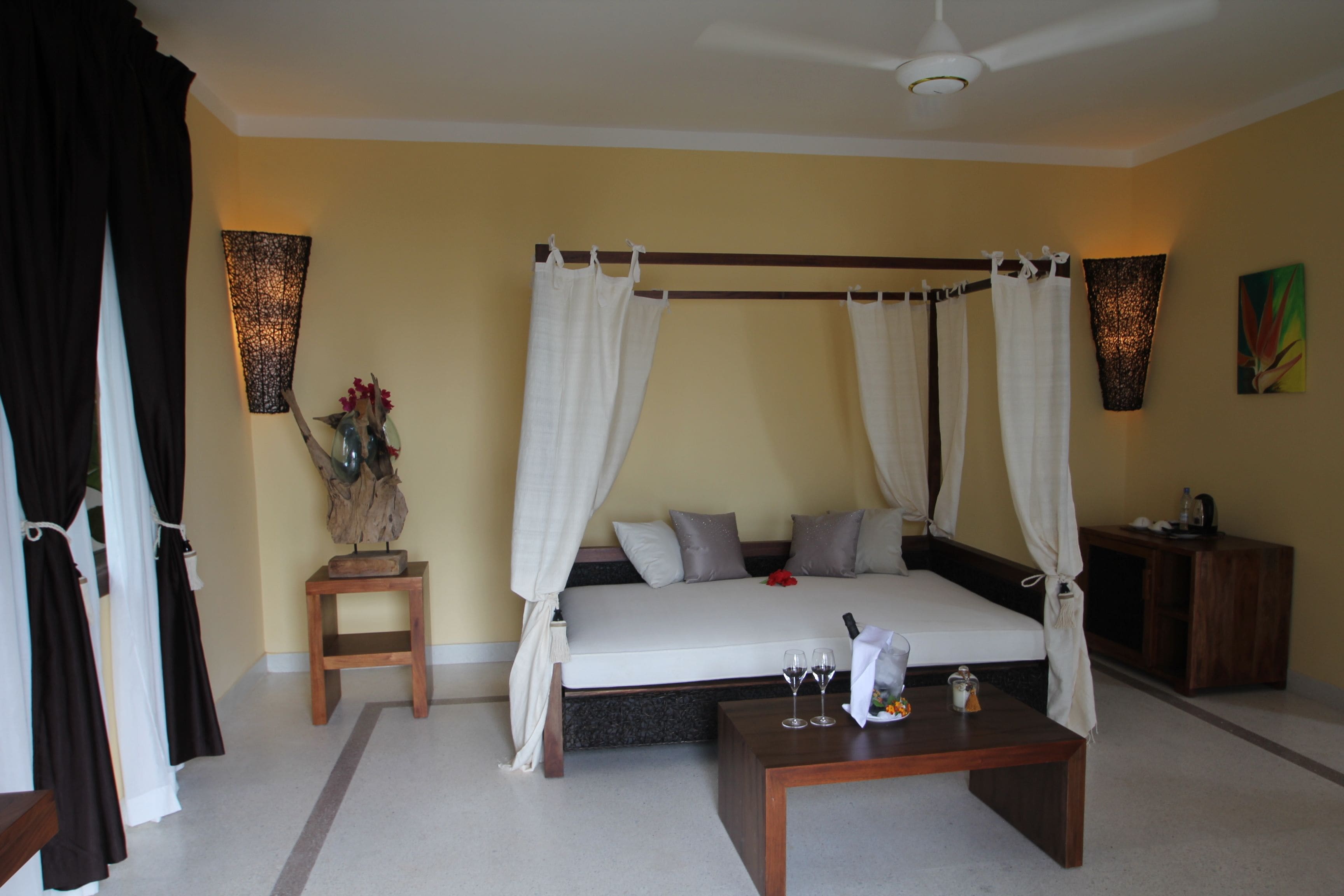A four-poster canopy bed with a wooden headboard, a bedside table with a vase of flowers, and a small wooden nightstand with a lamp.
