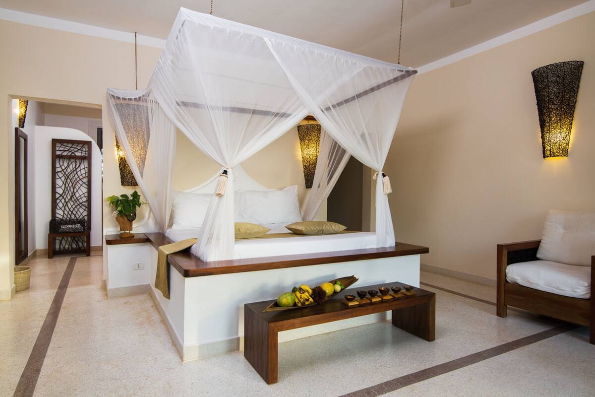 A bedroom with a canopy bed, a wooden bench with fruits, a small table, and a chair.