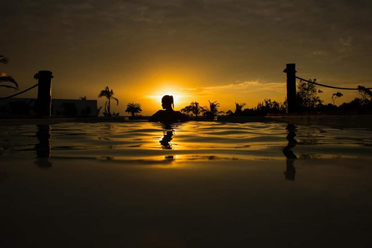 A silhouette of a person in a swimming pool at sunset with palm trees and a fence in the background.