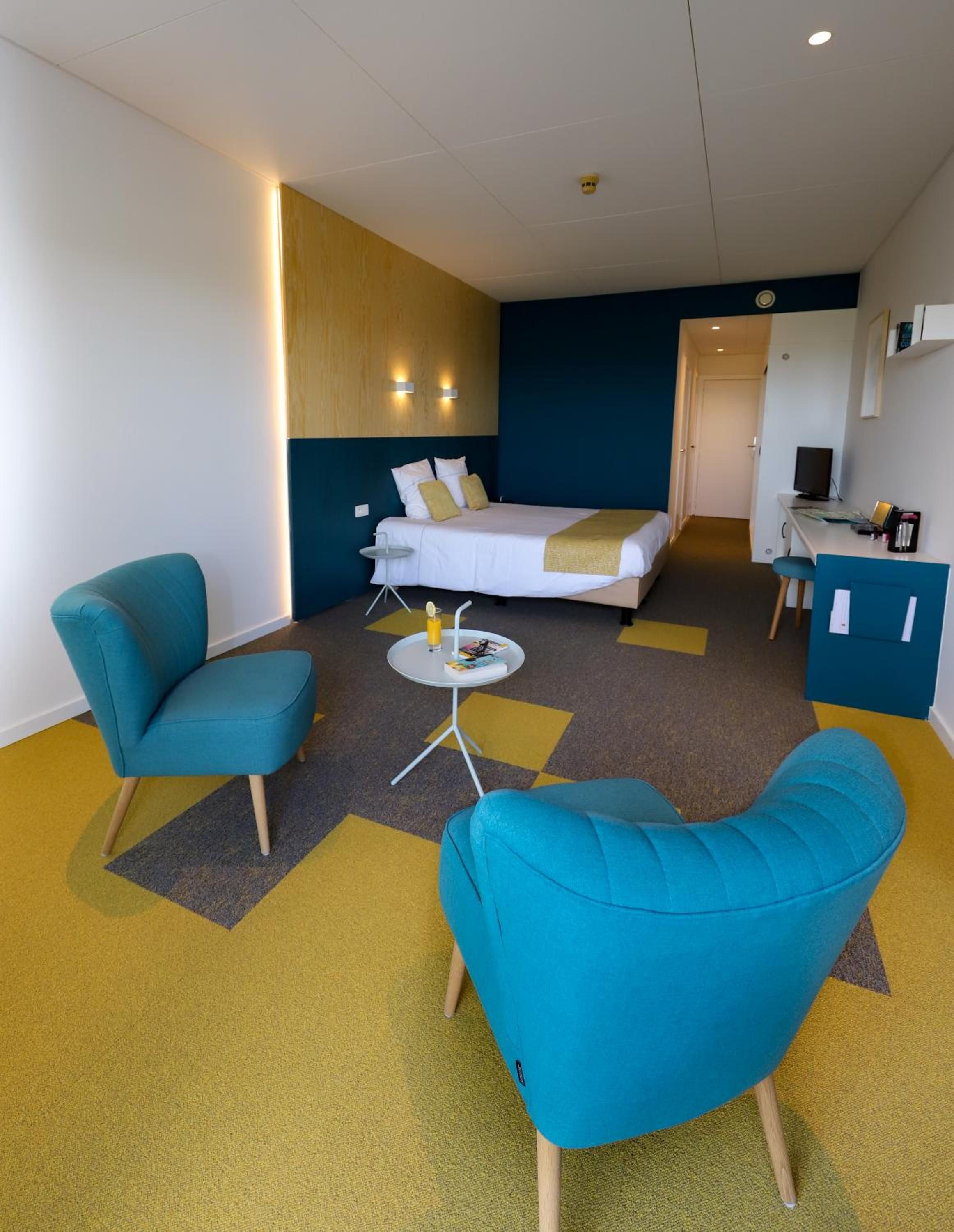 A hotel room with a bed, two blue armchairs, a small round table, and a desk with a computer.