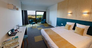 Una habitación de hotel con una cama grande, un escritorio con una computadora portátil, una silla y vistas al campo a través de un ventanal amplio.