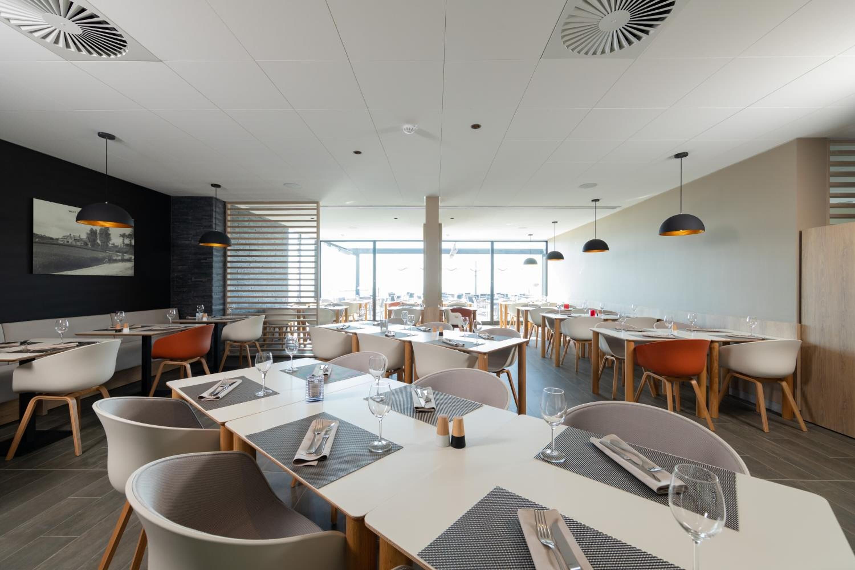 A modern dining area with multiple tables and chairs, large windows, and pendant lighting.