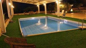 Una piscina rectangular con una superficie de agua azul cristalina, rodeada de césped artificial verde. Hay sillones de descanso y una silla de mimbre cerca de la piscina.