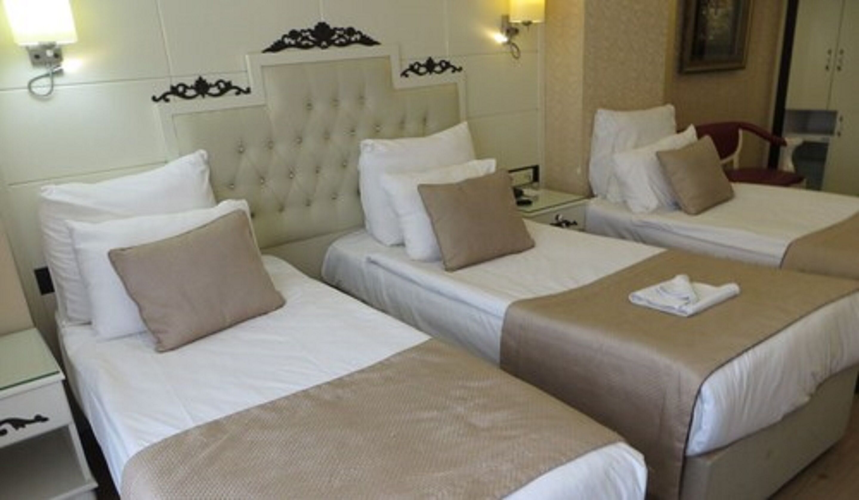 A hotel room with three single beds, each with a white duvet and a beige throw blanket. There are wall-mounted light fixtures and a framed picture above the beds.