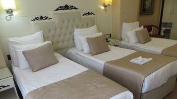 A hotel room with three single beds, each with a white duvet and a beige throw blanket. There are wall-mounted light fixtures and a framed picture above the beds.