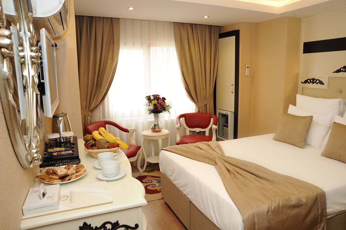 A hotel room with a bed, a desk with a coffee machine, a chair, and a small table with a vase of flowers.
