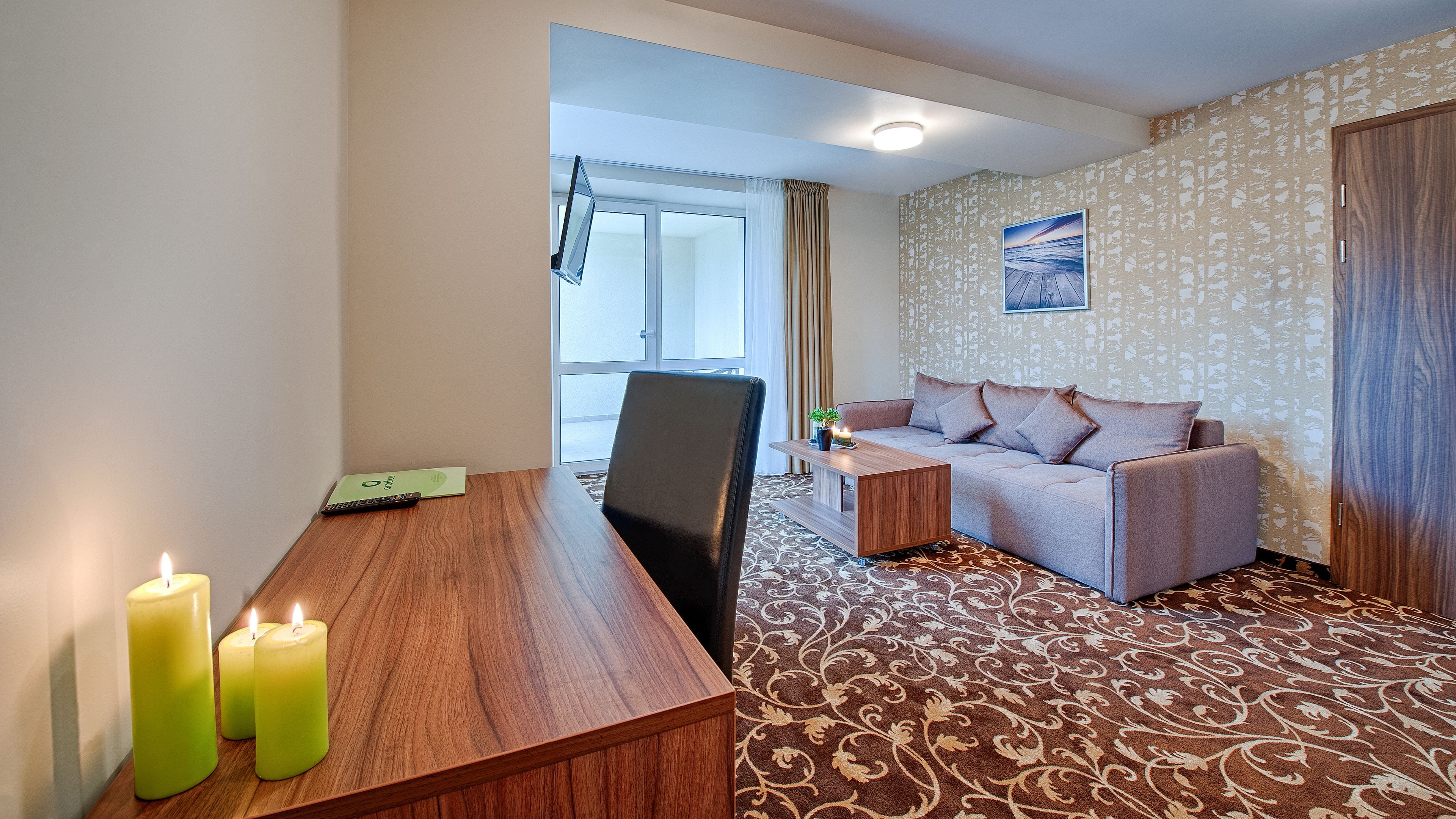 A hotel room with a wooden desk, two green candles, a grey sofa, and a patterned carpet.