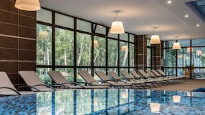 Una piscina interior moderna con paredes de vidrio transparente, sillas de descanso e iluminación colgante.