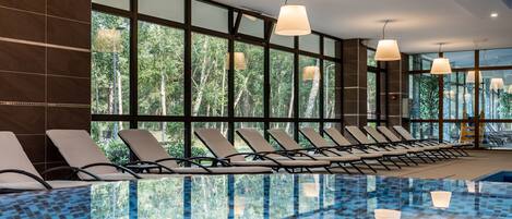 A modern indoor swimming pool with clear glass walls, lounge chairs, and pendant lighting.