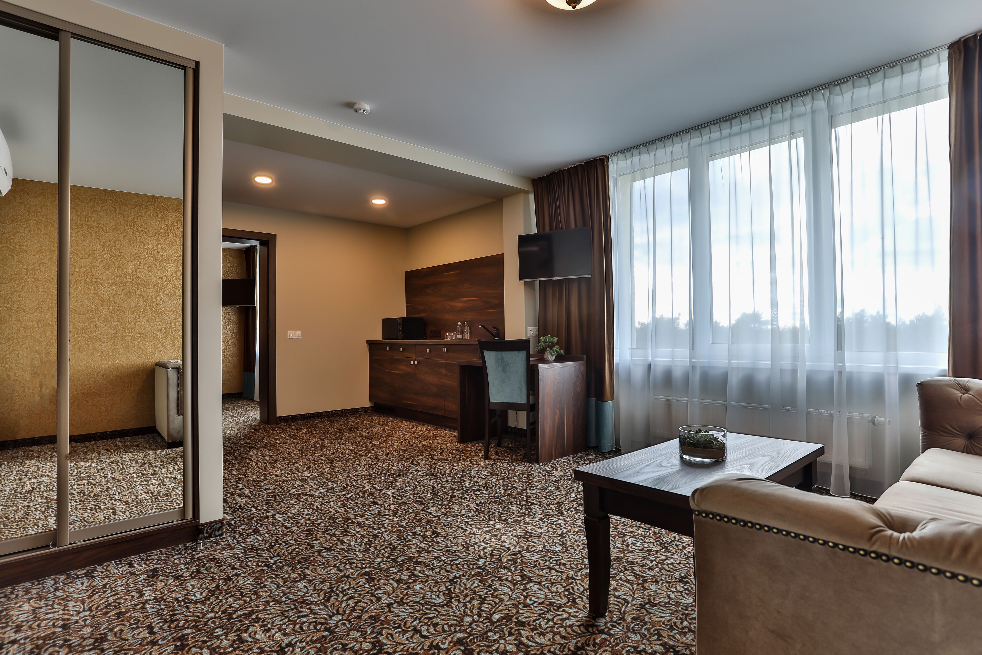 Habitación de hotel con alfombra estampada, un sofá, una mesa de centro, un televisor y vista al exterior a través de cortinas translúcidas.