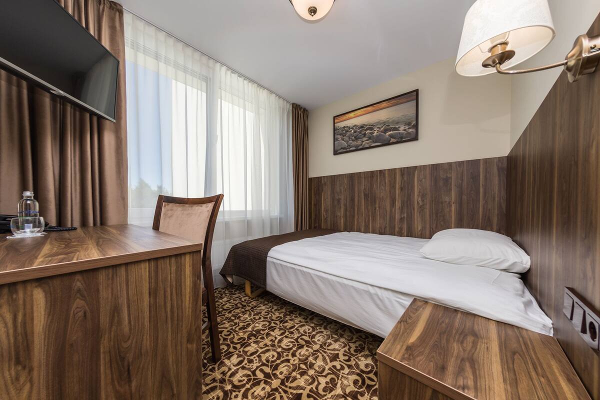 A hotel room with a bed, a desk, a chair, a TV, and a painting on the wall.