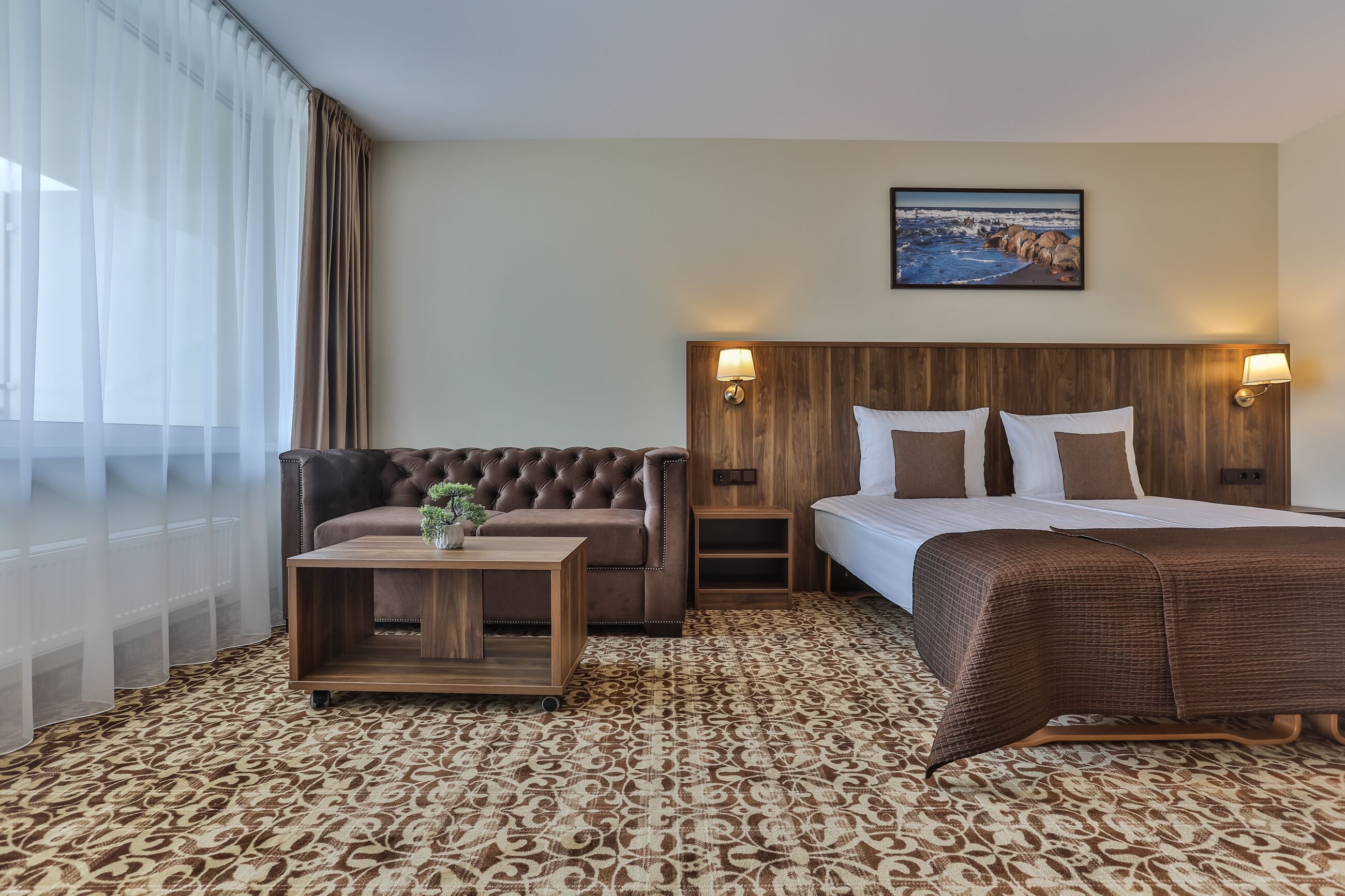 A hotel room with a large bed, a sofa, a small table with a plant, and a painting on the wall.