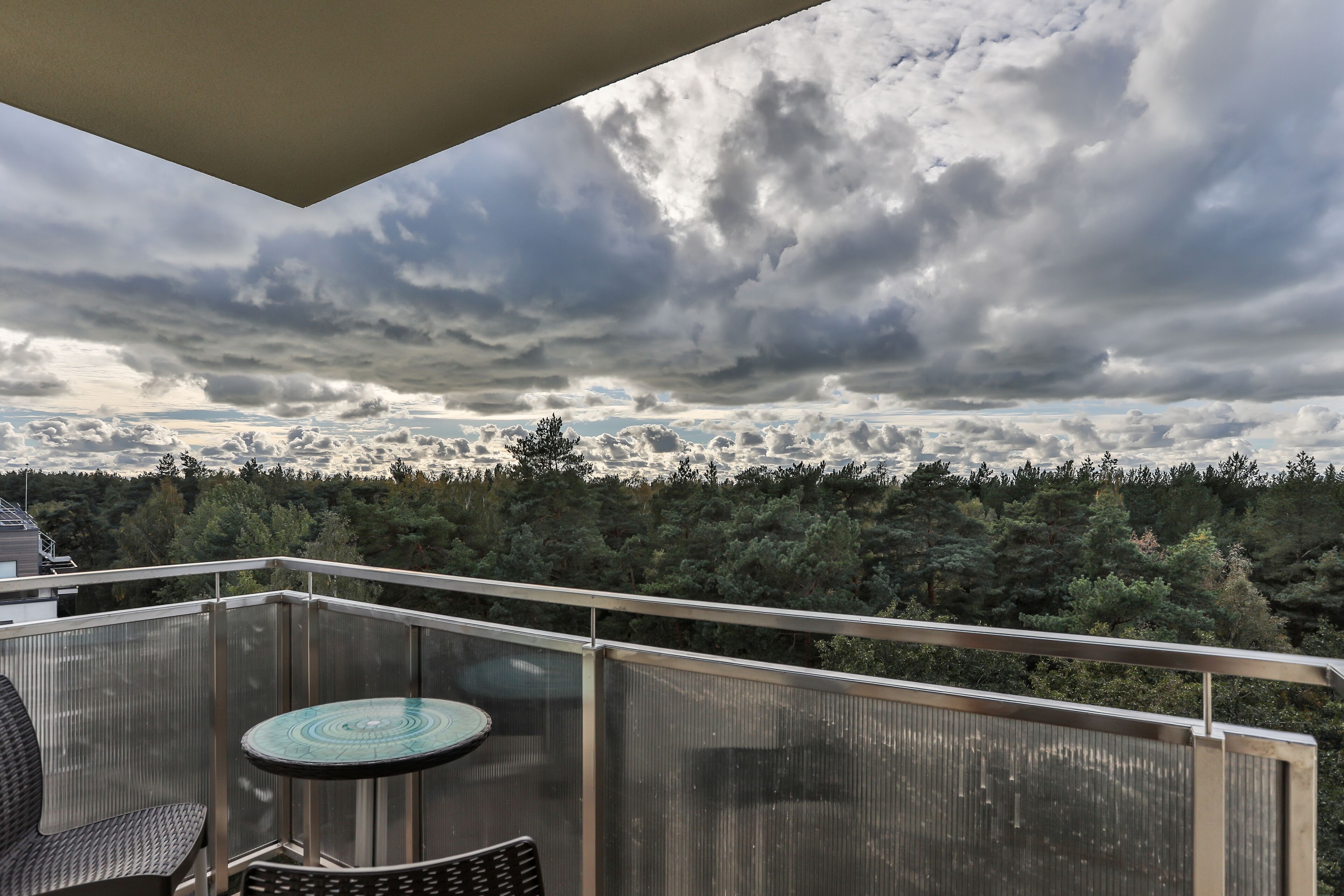 A balcony with a glass table and chairs, overlooking a forested area and a cloudy sky.