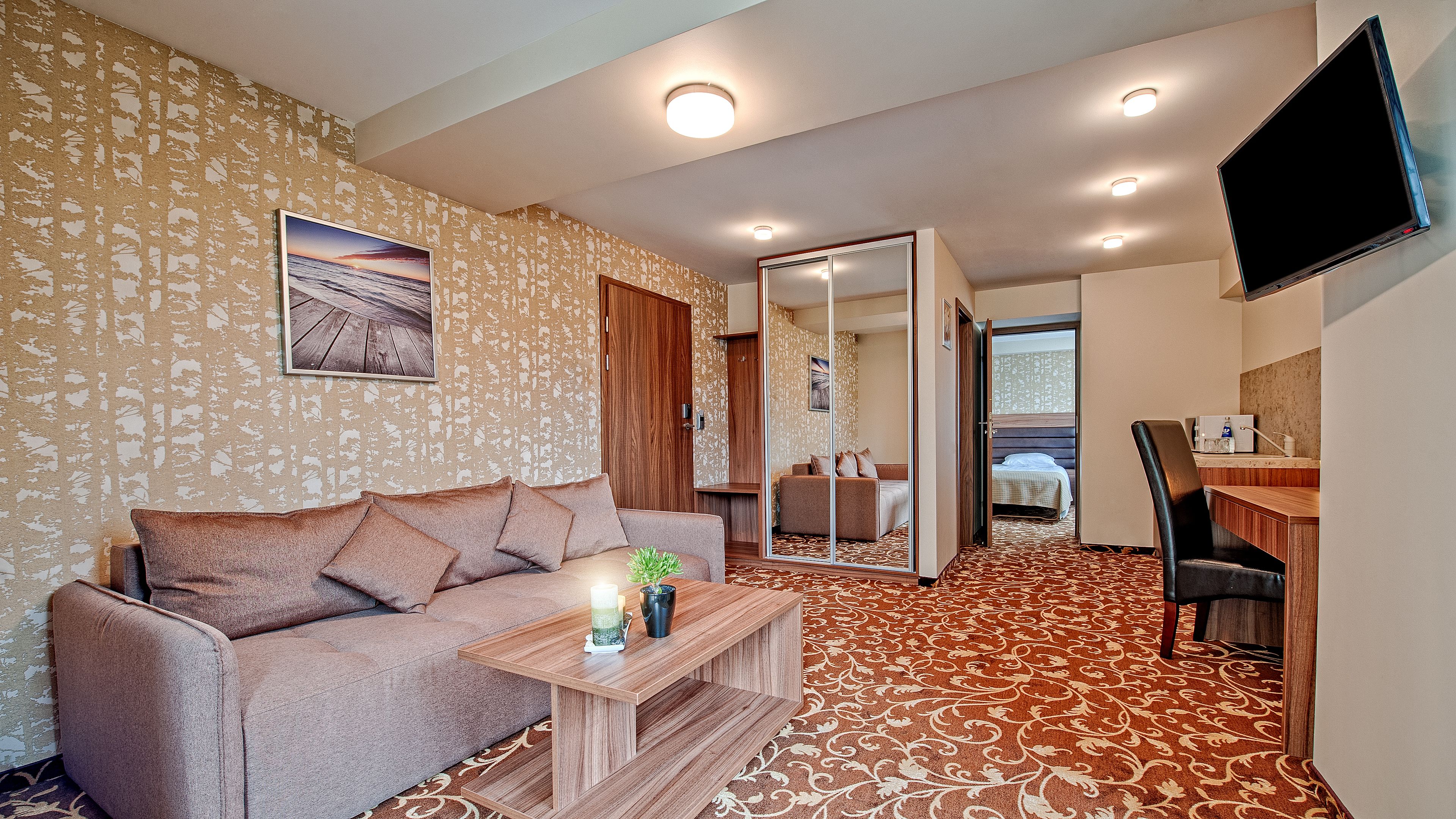 Una habitación de hotel moderna con un sofá, una mesita de madera, un televisor montado en la pared y una alfombra estampada.