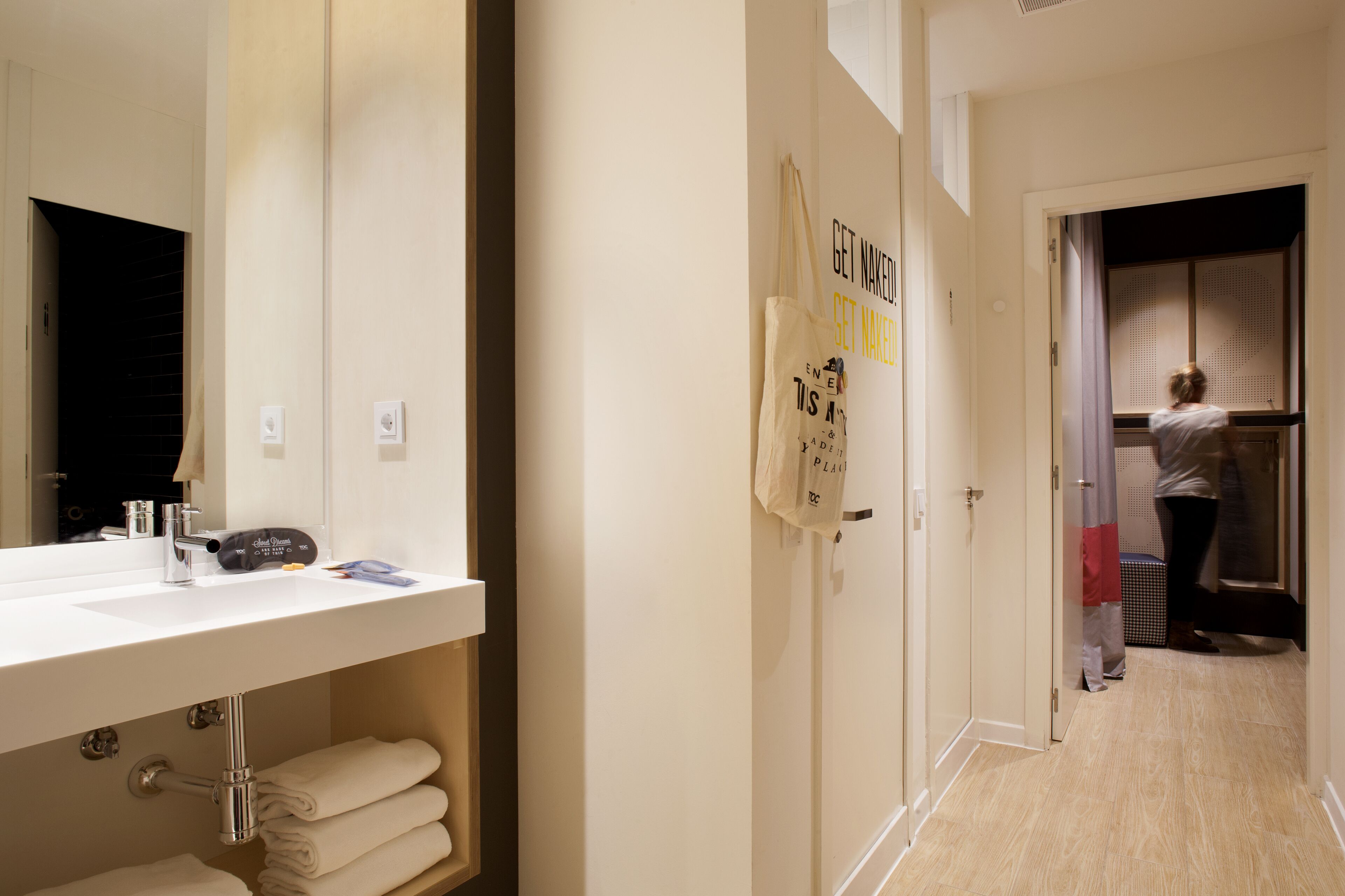 Un baño de hotel con lavamanos, toallero y espejo. Se ve una puerta que da a un armario con ropa colgada.
